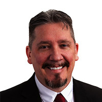 Headshot of a smiling person with dark hair and a goatee, wearing a dark suit and red tie